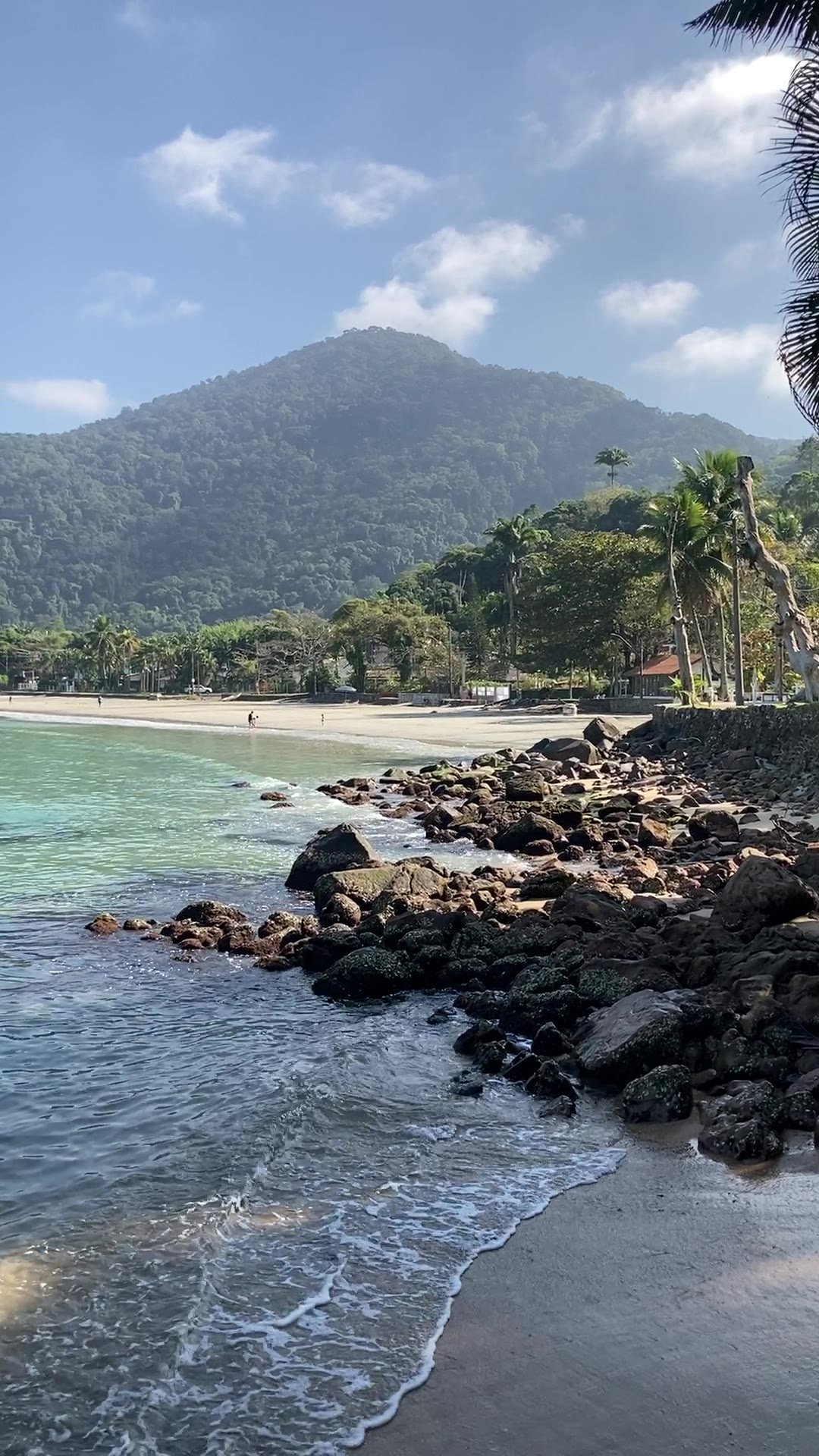 Orla da Praia da Enseada com pedras em primeiro plano e morro ao fundo