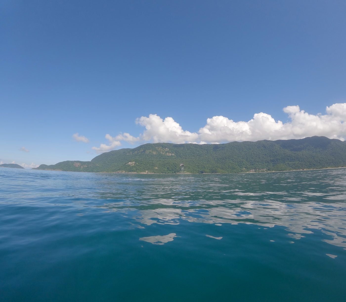 Mar aberto e liso com a costa de mata e nuvens ao fundo, visto do barco