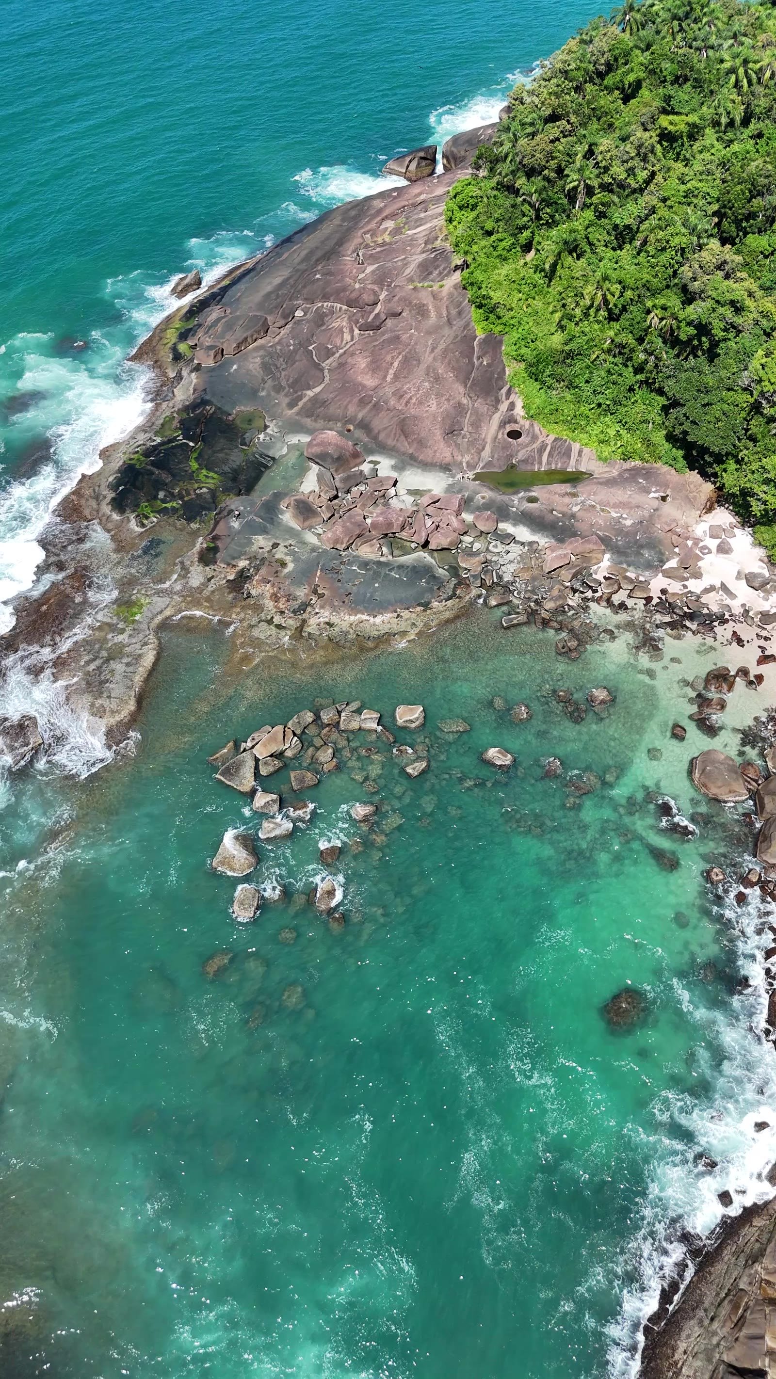 Vista de drone da Praia das Conchas em Ubatuba: costão de pedra, mata e água verde formando piscina entre as rochas