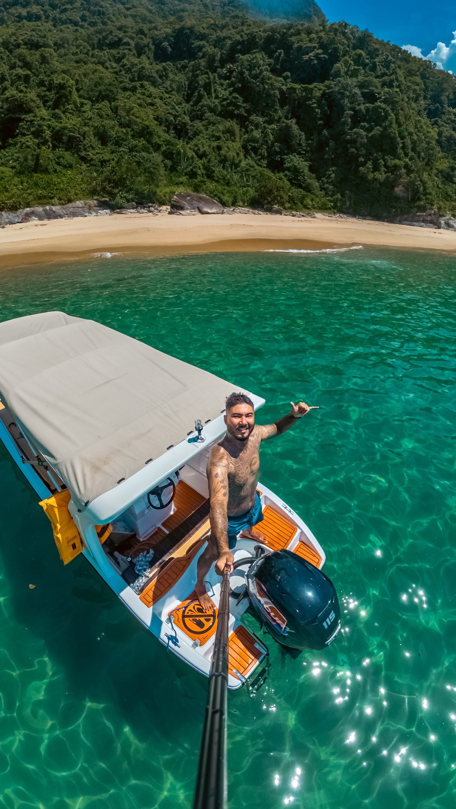 Renan em pé num barco sobre água verde e transparente, apontando pra praia